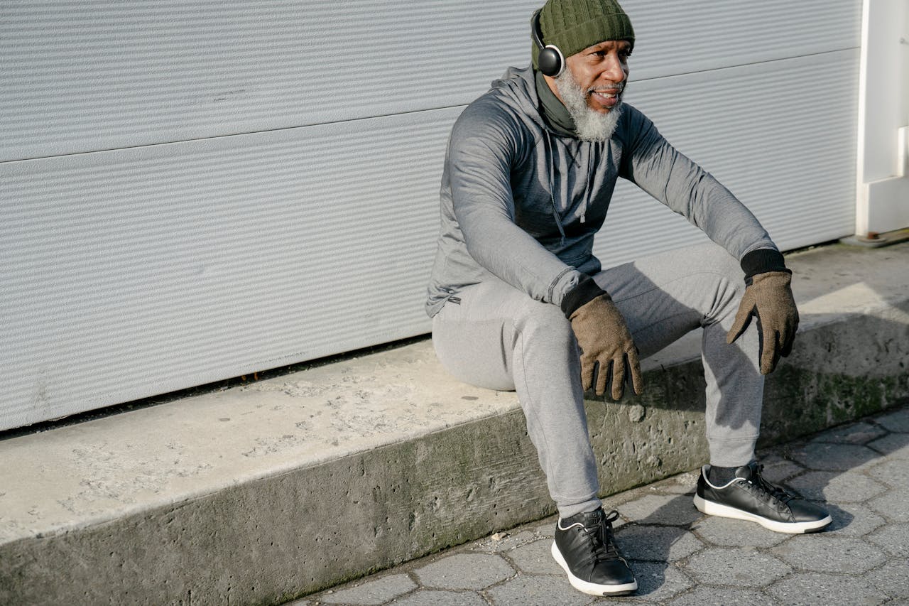 Full body of smiling bearded senior African American male wearing warm sportswear knitted hat and gloves sitting on curb of street and listening to music in headphones